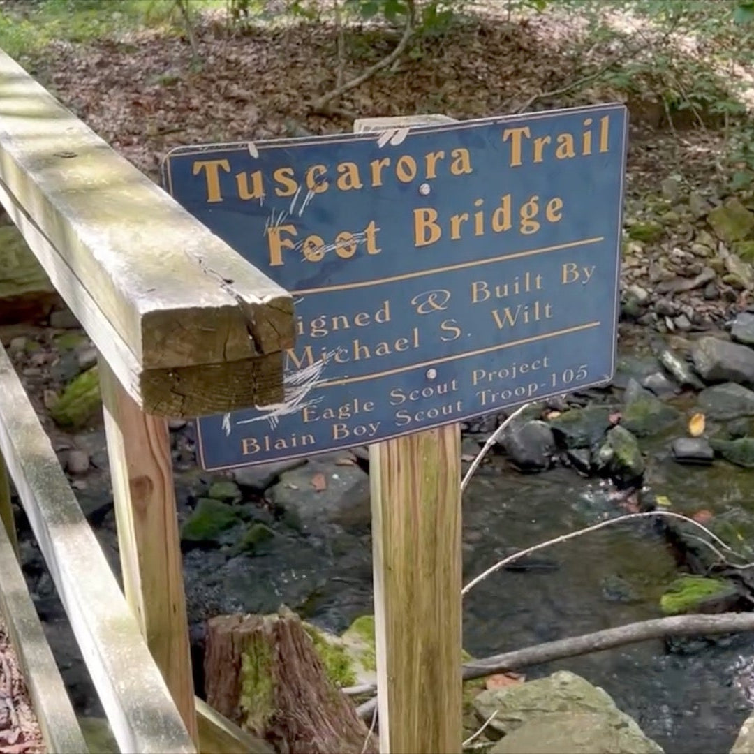 Wooden footbridge over a stream with a blue sign indicating 'Tuscarora Trail' in a forest setting.