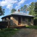Stone cabin with a wooden deck surrounded by trees on a sunny day.